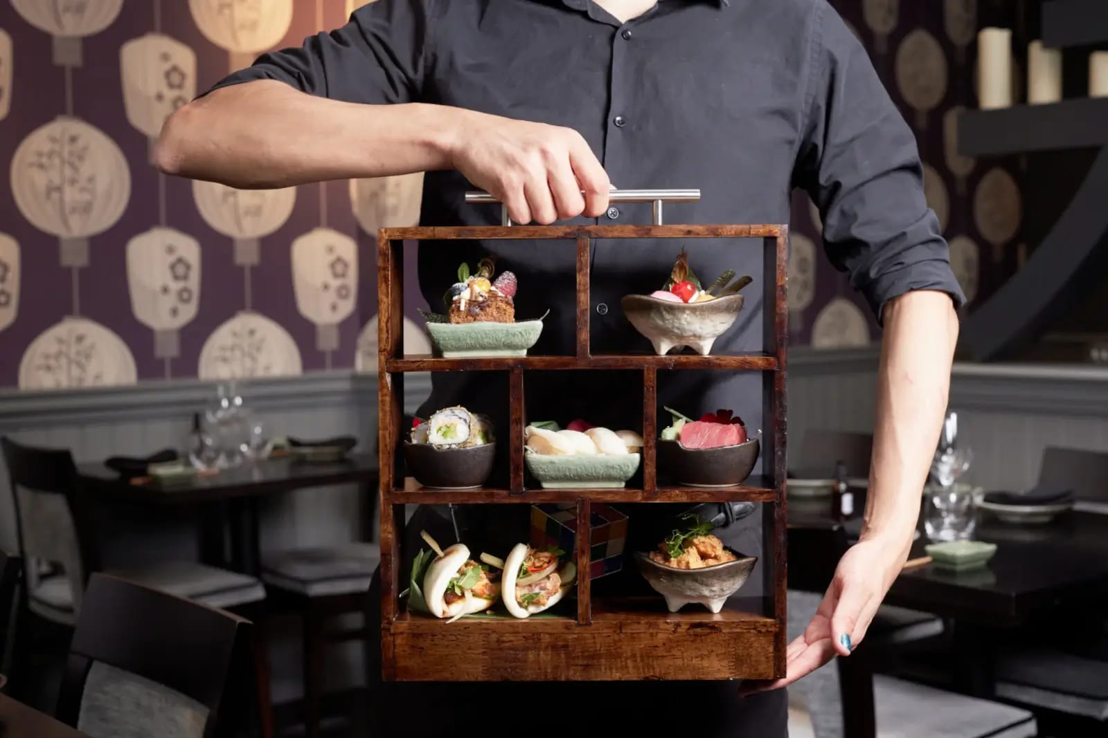 A server holding the wooden afternoon-tea cabinet, its pigeonholes filled with sushi, bao buns and desserts
